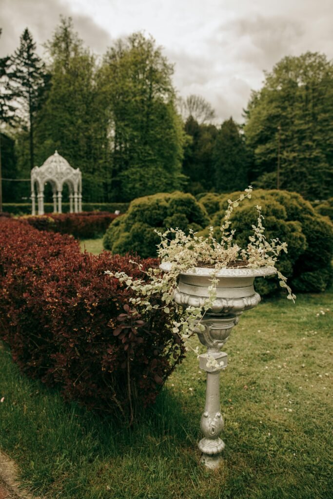 A picturesque garden with a vintage vase, lush greenery, and a distant gazebo under cloudy skies.